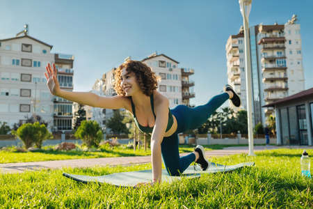 Young happy redhead woman wearing sportive clothes on yoga mat on city park, outdoors practicing...