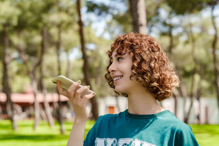 Young happy redhead woman wearing green tee standing on city park, outdoors using her mobile on speaker phone holding the device in front of face. Happy sportive woman talking with friends on phone.の写真素材