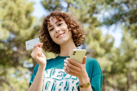 Young redhead woman wearing green t-shirt standing on city park, outdoors showing plastic credit or...