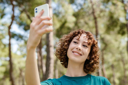 Cheerful woman wearing green casual clothes and standing on city park, outdoor taking a self portrait with smart phone. She looks at the screen and taking selfie.の写真素材