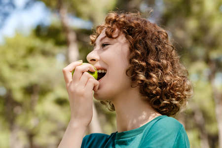 Side view of young happy redhead woman wearing green t-shirt standing on city park, outdoors biting fresh apple. healthy lifestyle. healthy eating. Fruits and vegetables.の写真素材