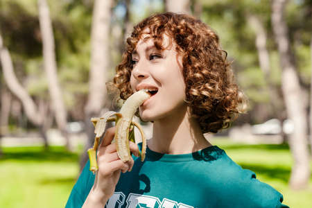 Happy young sportive woman wearing green t-shirt standing on city park, outdoors biting healthy...