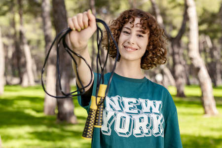 Cheerful redhead woman wearing green t-shirt standing on city park, outdoor holding her skipping rope in front of the camera. Outdoor sport, healthy life concepts.の写真素材