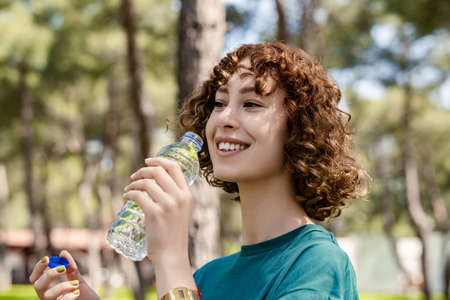 Close up view of happy redhead woman wearing green sportive clothes standing on city park, outdoors...