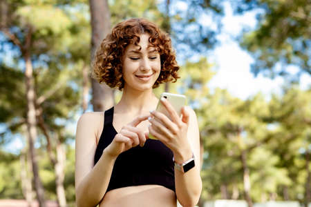 Happy redhead woman wearing black sports bra standing on city park, outdoors hands holding cell phone touching finger display, scrolling on social media, mobile application or tech concepts.の写真素材