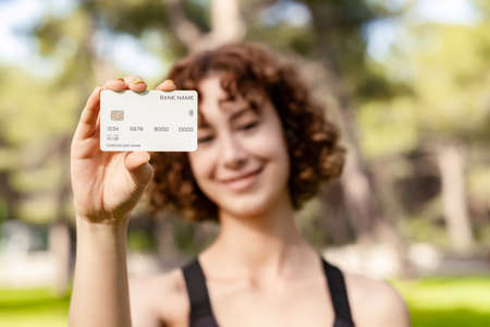 Young woman smiling confident wearing sportive clothes on city park, outdoors showing plastic credit or debit card, making online payments. Selective focus on credit card.の写真素材