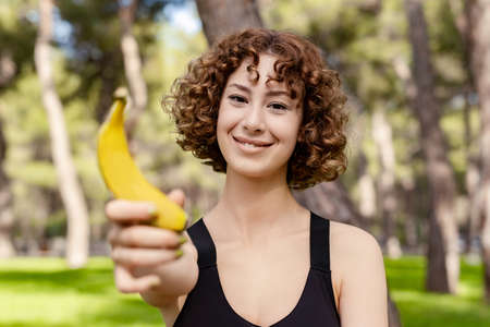 Young redhead woman wearing black sports bra standing on city park, outdoor holding a healthy banana and looking at the camera with smiley look. Outdoor sports and healthy life concepts.の写真素材