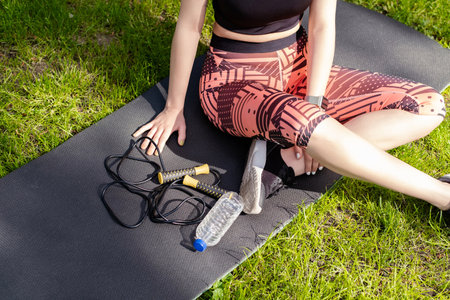 Young redhead woman wearing black sports bra standing on city park, outdoor sitting on a yoga mat...
