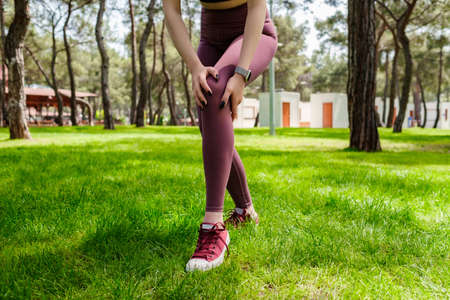 Brunette sportive woman wearing black sports bra standing on city park, outdoors touching injured knee with hands. Injured leg, healthy lifestyle and sport concept.の写真素材