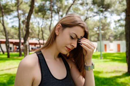 Young caucasian woman wearing black sports bra standing on city park, outdoors rubbing nose and eyes...
