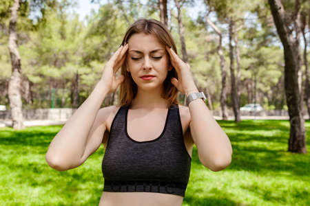 Young beautiful woman wearing sportive clothes on city park, outdoors holding her head with hands...