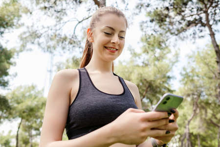 Cheerful brunette woman wearing sportive clothes on city park, outdoors looking at the phone screen and using phone. Messaging with friends, watching video or scrolling on social media.の写真素材