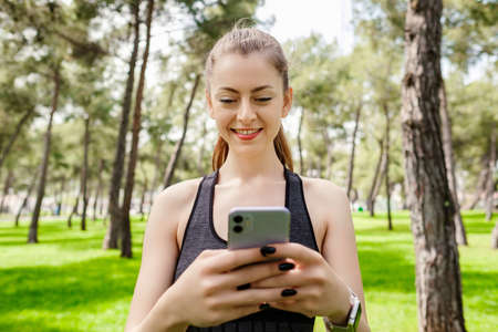 Happy sportive woman wearing sports bra standing on city park, outdoors looking at the phone screen...