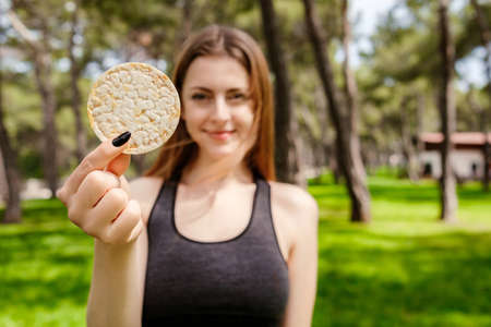 Cute sportive woman wearing black sports bra standing on city park, outdoors looking at the camera eating healthy rice crackers smiling happy and positive. Selective focus on rice crackers.の写真素材