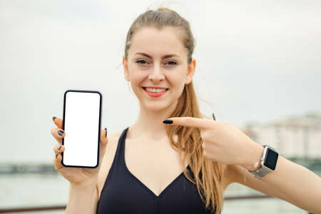 Happy brunette woman wearing black sports bra standing on city park, outdoors holding a phone and pointing finger to phone with smiley face. She is looking to cell phone screen.の写真素材