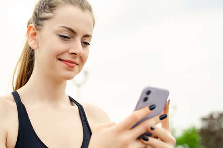 Cheerful brunette woman wearing sportive clothes on city park, outdoors looking at the phone screen and using phone. Messaging with friends, watching video or scrolling on social media.の写真素材