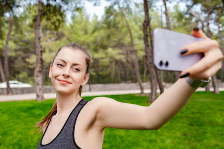 Young woman smiling confident wearing sports bra standing on city park, outdoors taking a self portrait with smart phone. She looks at the screen and taking selfie.の写真素材