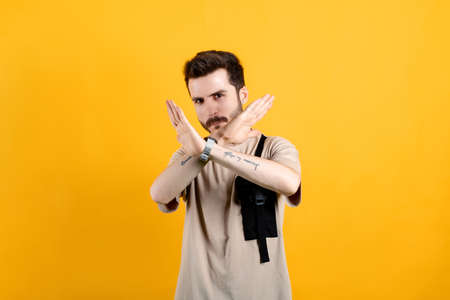 Young man wearing beige t-shirt posing isolated over yellow background rejection expression crossing...