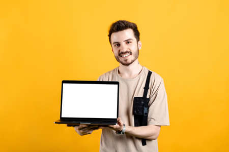 Young man smiling confident wearing beige tee posing isolated over yellow background holding laptop pc computer with blank empty screen.の写真素材