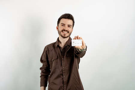 Cheerful young man wearing brown shirt posing isolated over white background showing his plastic credit or debit card with both hands. Shopping and finance concept.の写真素材