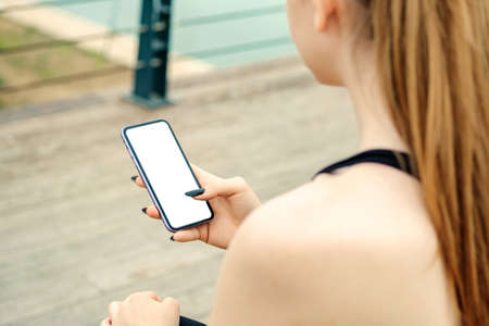 Brunette sportive woman wearing black sports bra standing on city park, outdoors hands holding phone touching finger mockup white blank display, mobile application tech concept, over shoulder view.の写真素材