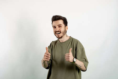 Cheerful young man wearing t-shirt posing isolated over white background smiling and showing thumbs up at camera. Approving expression looking at the camera showing success.の写真素材