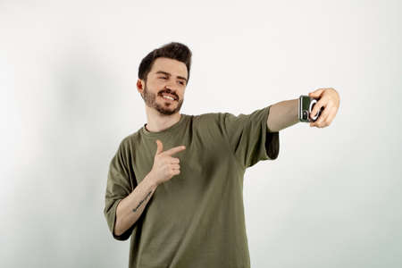Young man wearing khaki t-shirt posing isolated over white background pointing index finger on mobile phone while taking selfie. Communicating concepts.の写真素材