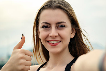 Cute sportive woman wearing black sports bra standing on city park, outdoors showing thumb up and...