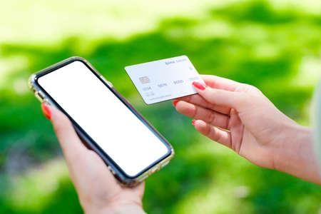 Brunette millennial woman wearing turquoise tee on city park, outdoors holding blank screen mobile phone credit bank card do online shopping provide internet payment. Safe mobile banking.の写真素材