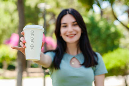 Cheerful brunette woman wearing tee standing on city park, outdoors showing takeaway cup of coffee. Selective focus on coffee mug.の写真素材