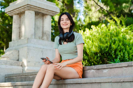 Happy brunette woman wearing turquoise tee on city park, outdoors sitting on stairs and looking at...