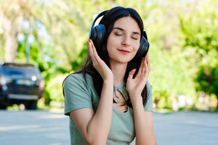 Happy brunette woman wearing turquoise tee on city park, outdoors listening music in headphones and...