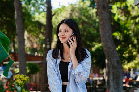 Young brunette woman wearing blue striped shirt on city park, outdoors talking on mobile phone with friends or boyfriend with smiles.の写真素材