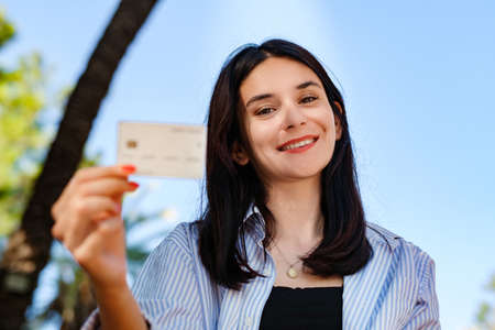 Cute caucasian woman wearing striped shirt standing on city park, outdoors showing plastic credit or debit card while holding mobile phone. Online shopping, banking and transaction concepts.の写真素材