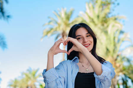 Cute brunette woman wearing shirt standing on city park, outdoors showing heart gesture with two hands and looking at camera. Heart shape with hands.の写真素材