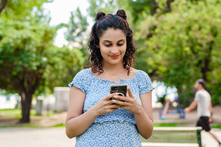 Portrait of brunette woman wearing summer dress standing on city park, outdoors looking at the phone screen and using phone. Messaging with friends, watching video or scrolling on social media.の写真素材