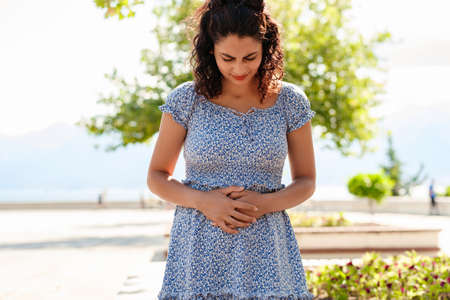 Young beautiful woman wearing summer dress on city park, outdoors with hand on stomach because...