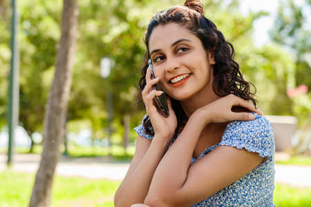 Young brunette girl smiling happy wearing summer dress on city park, outdoors talking on mobile phone with friends or boyfriend with smiles.の写真素材