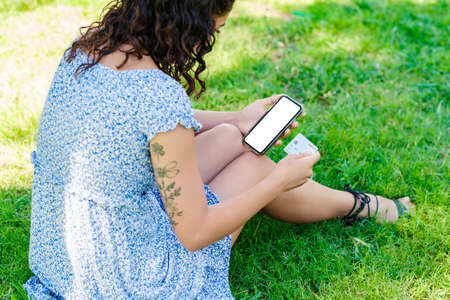 Cute brunette woman wearing summer dress standing on city park, outdoors holding credit card and phone with mockup white blank display, empty screen for shopping app. Over shoulder close up view.の写真素材