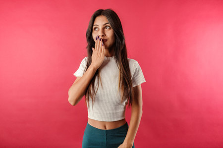 Young dark-haired woman wearing ribbed crop isolated over red background smiles looking at the copy space while closing mouth having heard the gossip. Giggle, hears gossip, looks amazed by rumour.の写真素材