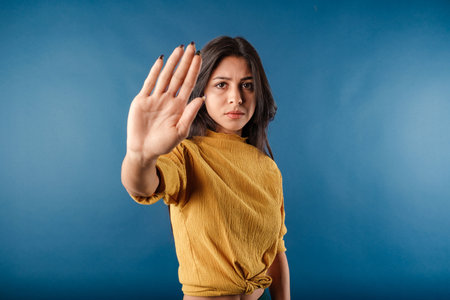 Portrait of brunette woman wearing mustard yellow t-shirt isolated over blue background doing stop...