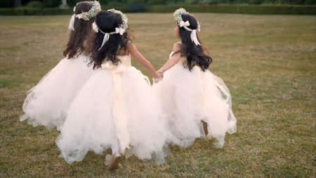 Back view of two beautiful bridesmaids in white dresses and wreaths holding hands while walking in the park.の写真素材