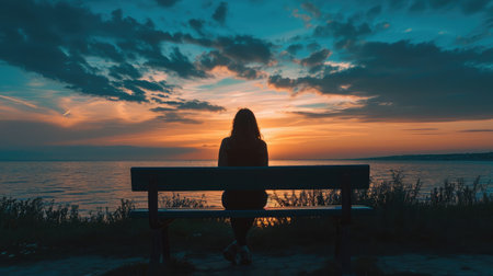 Wide angle shot of the Silhouette of a woman sitting on a bench looking at the tranquil sunset over the oceanの素材