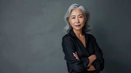 Stylish confident adult 50 years old Asian female psychologist standing arms crossed looking at camera at gray background.の素材