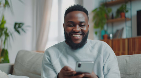 Smiling bearded African man working at home while sitting on the sofa.Concept of young people using mobile devices.Blurred background.Croppedの素材