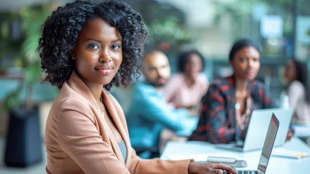 Attractive african young confident businesswoman sitting at the office table with group of colleagues in the background, working on laptop computerの素材