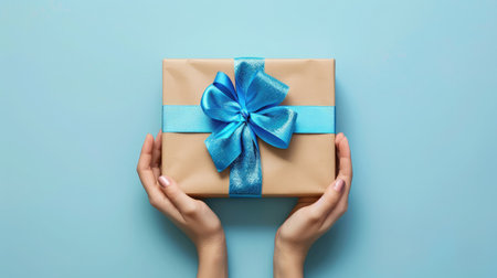 First person top view photo of hands holding craft paper giftbox with vivid blue ribbon bow over shiny sequins on isolated pastel blue backgroundの素材
