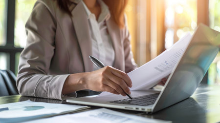 Serious focused businesswoman typing on laptop holding papers preparing report analyzing work results, female executiveの素材