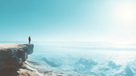 A serene moment captured of an individual standing on a rocky cliff, gazing into the expansive ocean under a brilliant blue sky with graceful waves.の素材