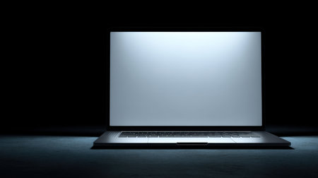 A sleek modern laptop sits in darkness with an illuminated blank screen, perfect for depicting themes of technology, minimalism, and online engagement in various settings.の素材
